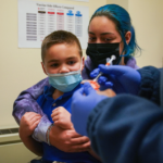 A woman holds a young boy while in the foreground a doctor's hands are visible preparing a needle.