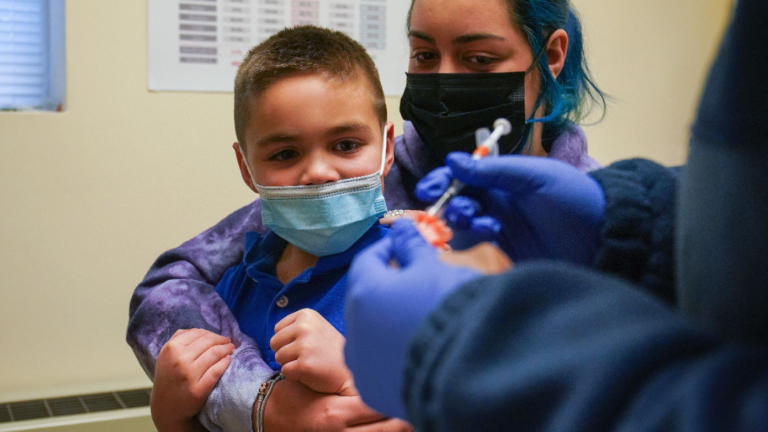 A woman holds a young boy while in the foreground a doctor's hands are visible preparing a needle.