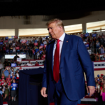 Donald Trump in the foreground in a dark blue suit and red tie. Behind him, a crowd of supporters with signs and Trump gear.