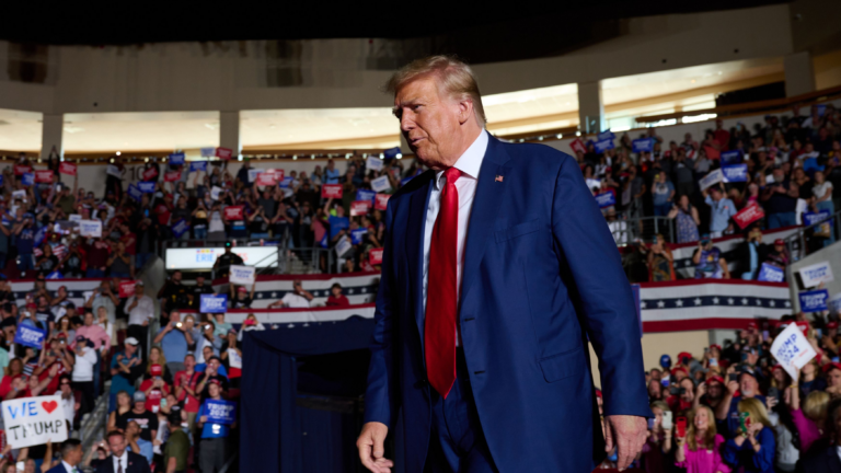 Donald Trump in the foreground in a dark blue suit and red tie. Behind him, a crowd of supporters with signs and Trump gear.