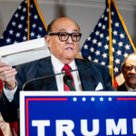 Giuliani, wearing a suit, speaks from a podium at a Trump campaign event with American flags in the background
