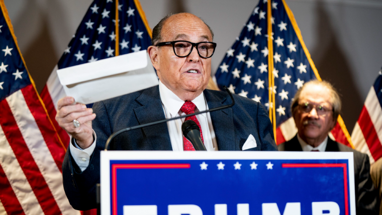 Giuliani, wearing a suit, speaks from a podium at a Trump campaign event with American flags in the background
