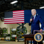Joe Biden speaks into a microphone at a table with the President's seal. Behind him is factory machinery and an American flag.