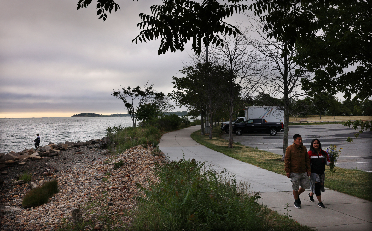 A couple walks along a paved path overlooking the Boston Harbor.