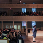 Men in Revolutionary-era clothing and wigs fill the interior of the Old South Meeting House. Most sit in pews. One stands before the crowd, speaking.