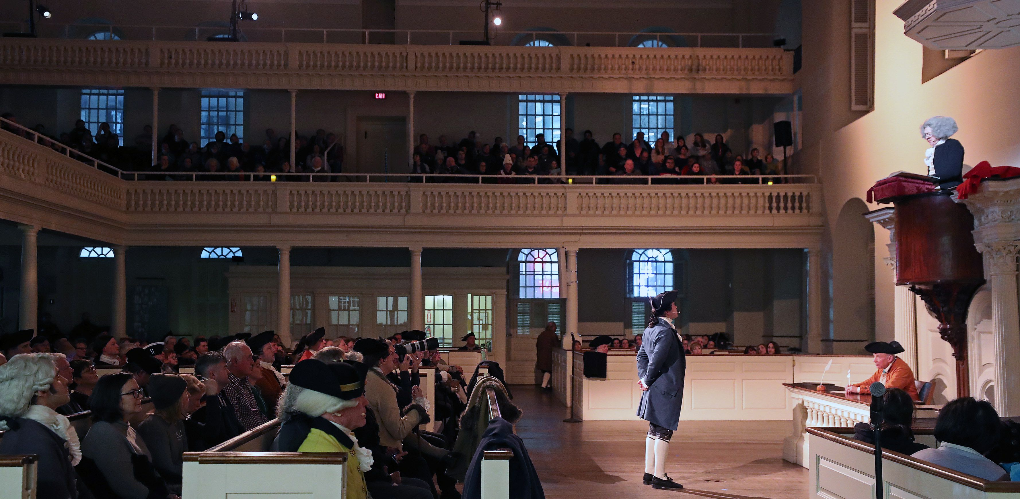 Men in Revolutionary-era clothing and wigs fill the interior of the Old South Meeting House. Most sit in pews. One stands before the crowd, speaking.