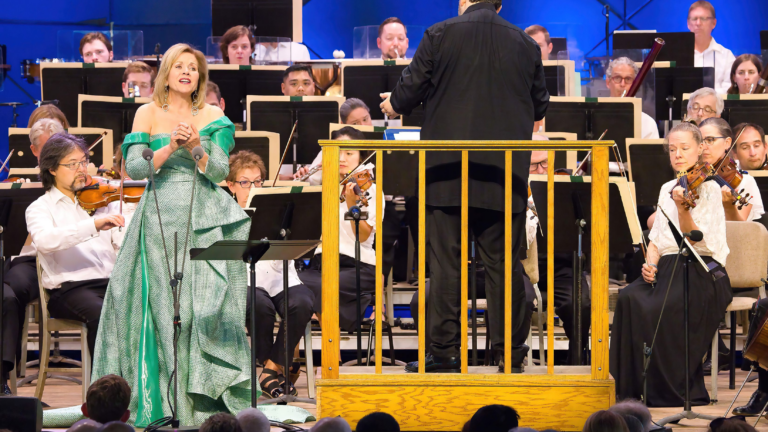 A woman in a teal blue gown sings in front of an orchestra.