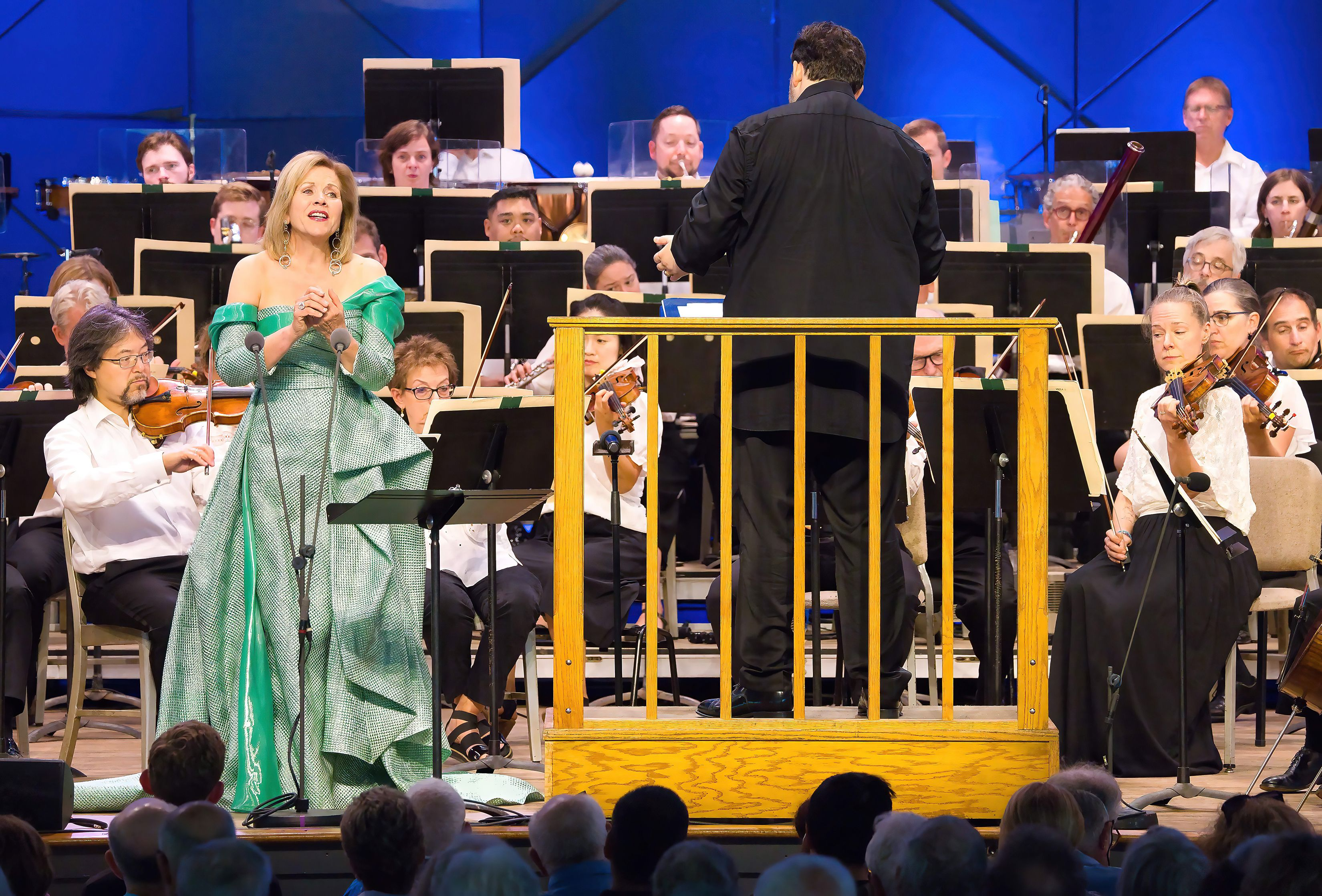 A woman in a teal blue gown sings in front of an orchestra.