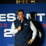 Ron DeSantis speaks into a microphone in front of a "DeSantis for President 2024" sign.