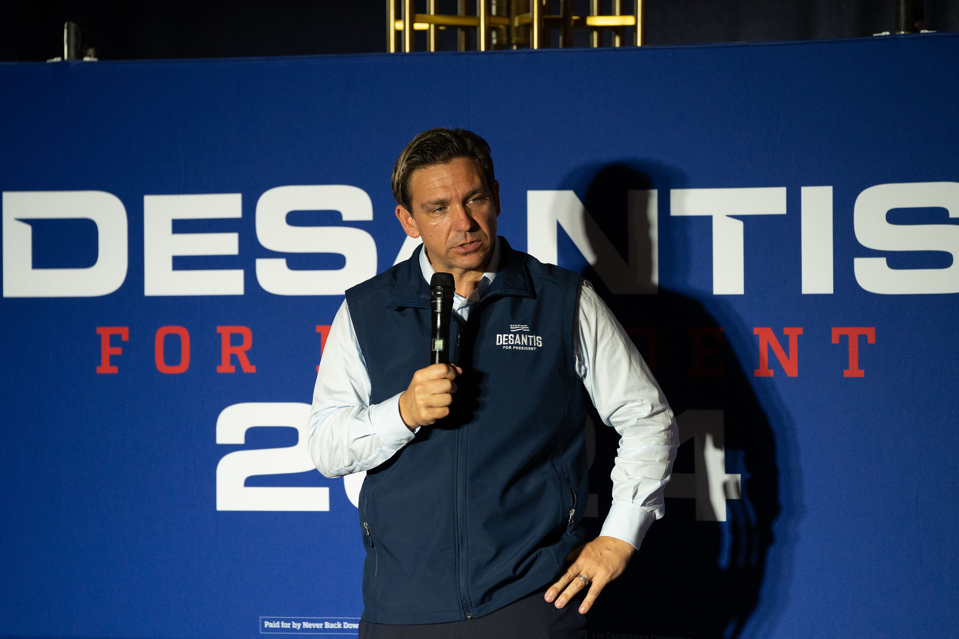 Ron DeSantis speaks into a microphone in front of a "DeSantis for President 2024" sign.