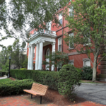 An academic building surrounded by trees and bushes, with a bench in the foreground