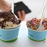 Two cups of ice cream are pictured side by side on a counter, one with chocolate jimmies and one with rainbow sprinkles.