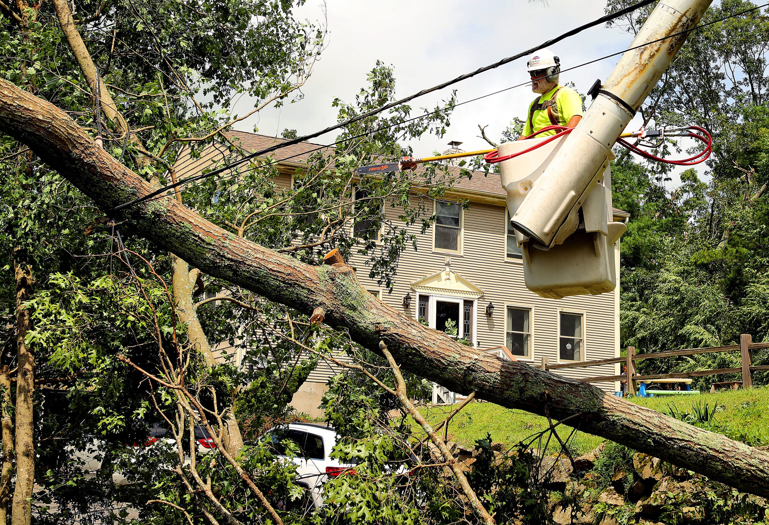 A worker in a bucket truck cuts branches on a downed tree that is atop power lines, in front of a brown two-story home.