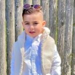 A 2-year-old boy in a white button-up shirt, cream-colored vest, khakis with circle-shaped sunglasses resting on top of his head poses for a picture outside in front of a wooden fence.