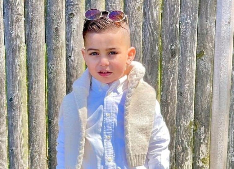 A 2-year-old boy in a white button-up shirt, cream-colored vest, khakis with circle-shaped sunglasses resting on top of his head poses for a picture outside in front of a wooden fence.
