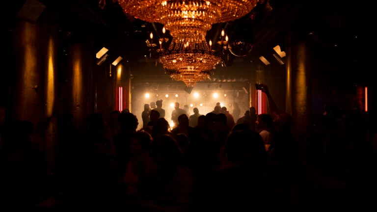A dim nightclub in Boston, showing silhouetted patrons in front of a stage. A chandelier hangs above the crowd.