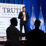 With a microphone in hand, Vivek Ramaswamy takes questions during a town hall while standing in front of a large white poster reading the word "Truth."