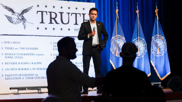 With a microphone in hand, Vivek Ramaswamy takes questions during a town hall while standing in front of a large white poster reading the word "Truth."
