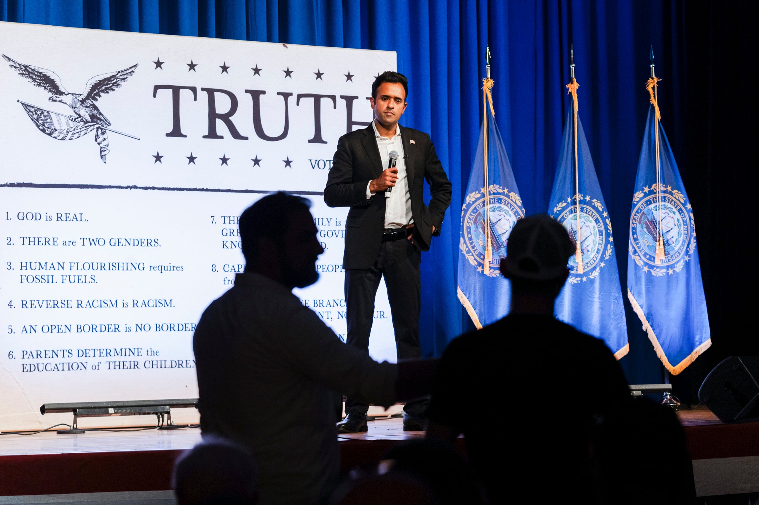 With a microphone in hand, Vivek Ramaswamy takes questions during a town hall while standing in front of a large white poster reading the word "Truth."