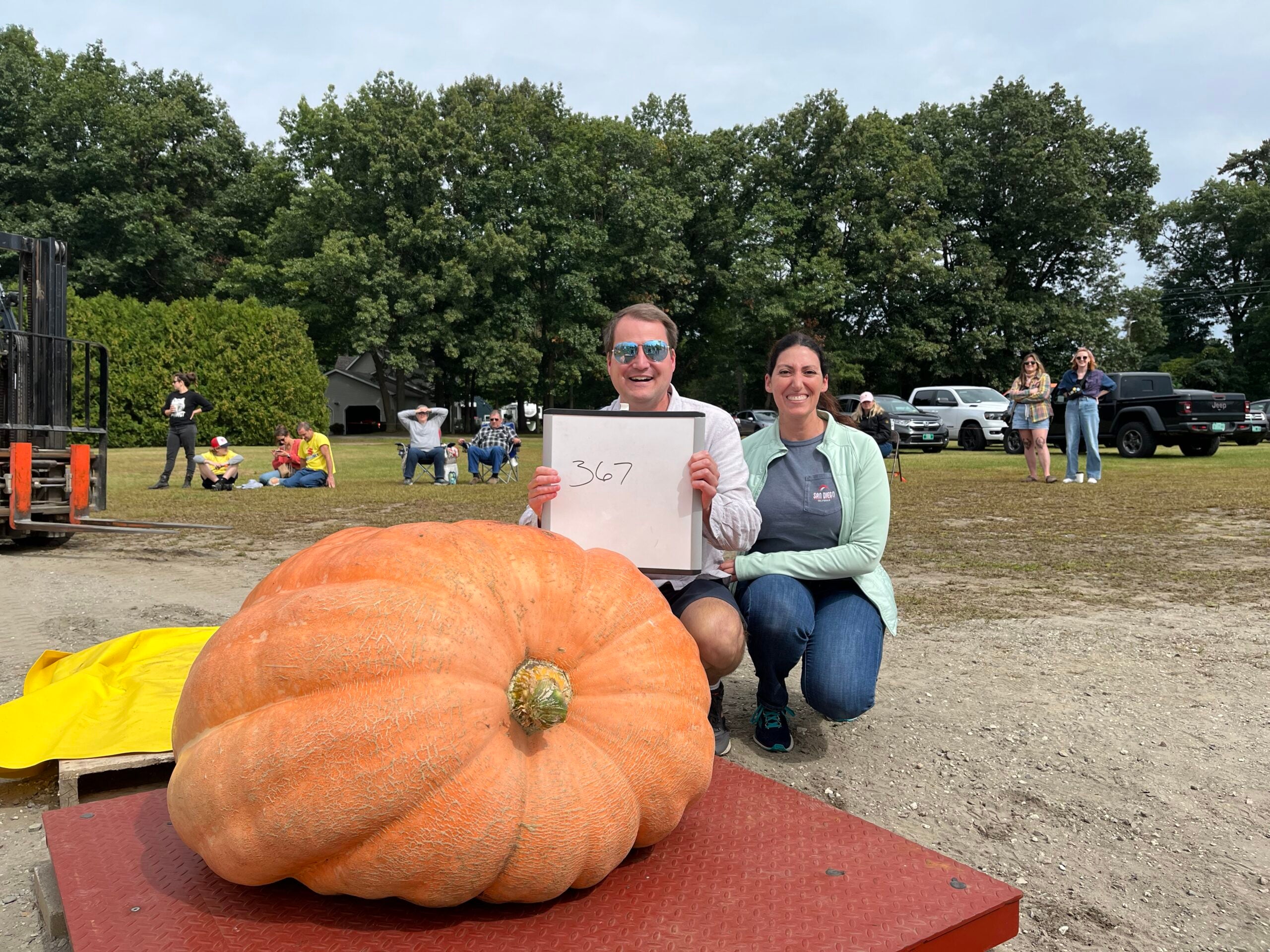 A man holds a sign that reads 367, the weight of an orange pumpkin in front of him on a scale. A woman sits next to him, both posing for a photo.