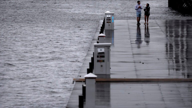 Two people are seen walking on a dock in the Seaport District of Boston along the water on a stormy day.
