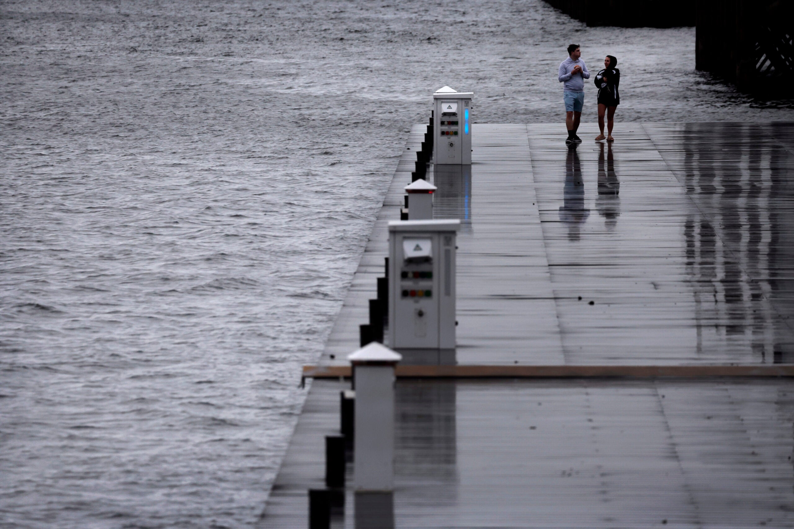 Two people are seen walking on a dock in the Seaport District of Boston along the water on a stormy day.