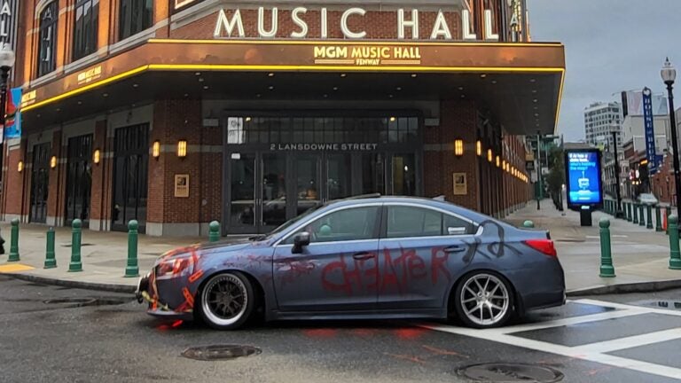 A picture of a gray Subaru, covered in caution tape and red paint that spells out the word "ch3ater." The car is parked in front of the MGM Music Hall in Boston.