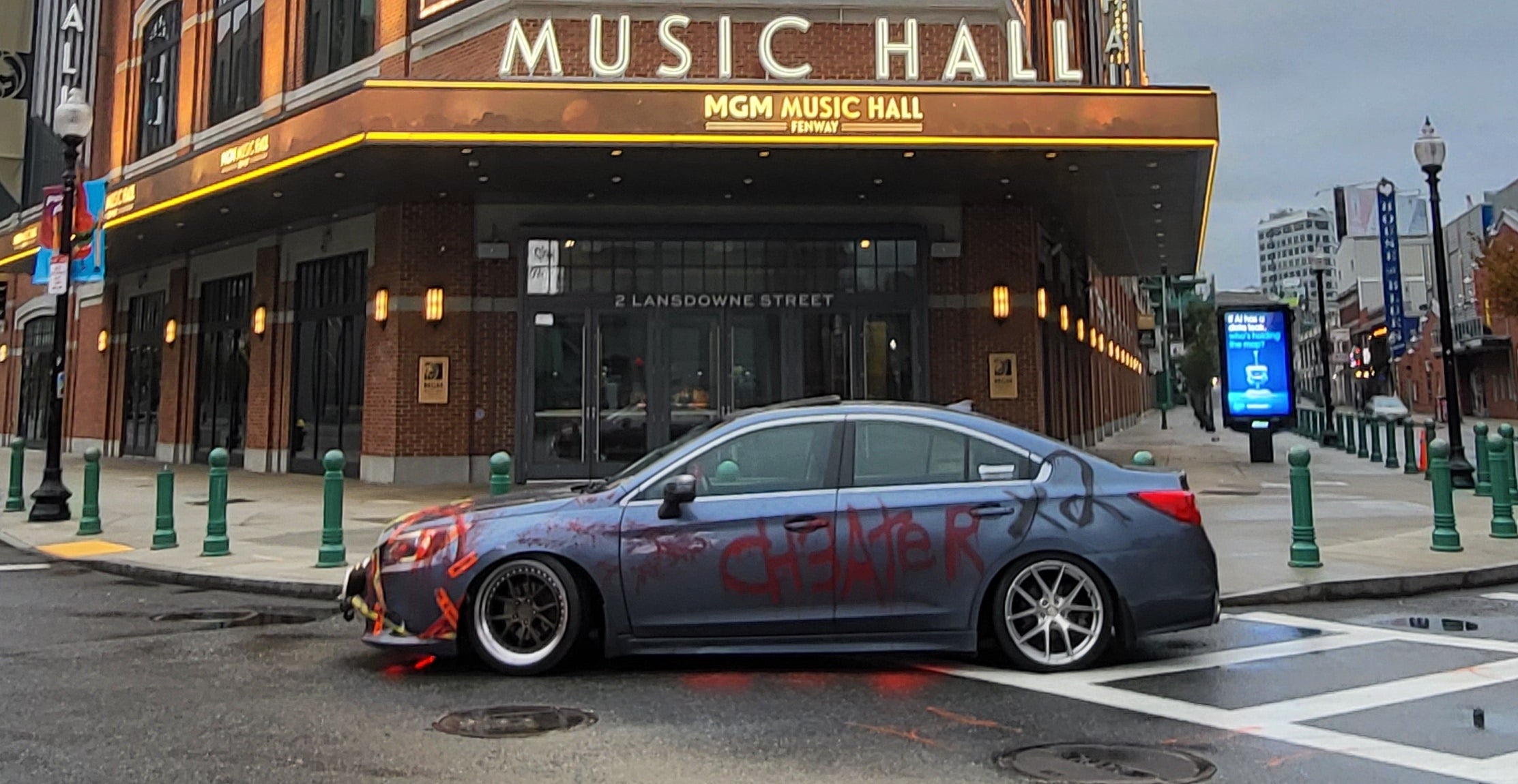 A picture of a gray Subaru, covered in caution tape and red paint that spells out the word "ch3ater." The car is parked in front of the MGM Music Hall in Boston.