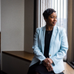 Andrea Campbell, seated wearing a blue blazer, gazes thoughtfully out a window
