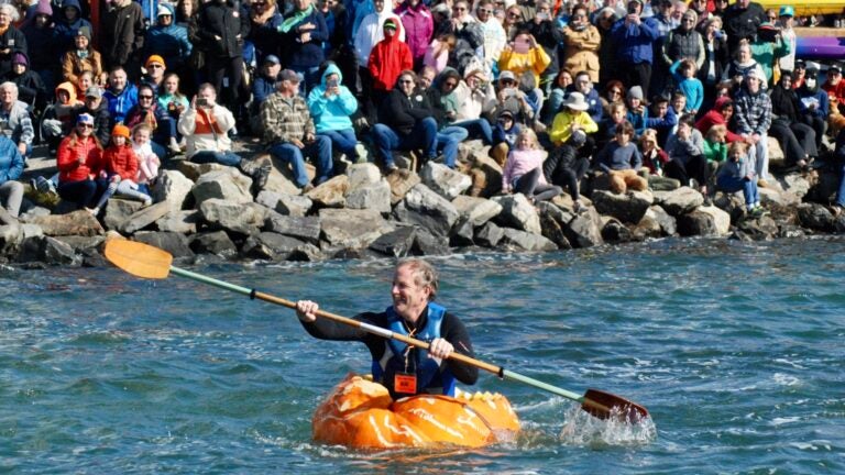 A man is seen paddling in a boat made out of a giant pumpkin in Maine, with a crowd of spectators behind him.