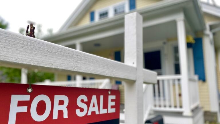 yellow home with blue shutters and a farmer's porch with a white railing has a for-sale sign out front that has white lettering on a red background.