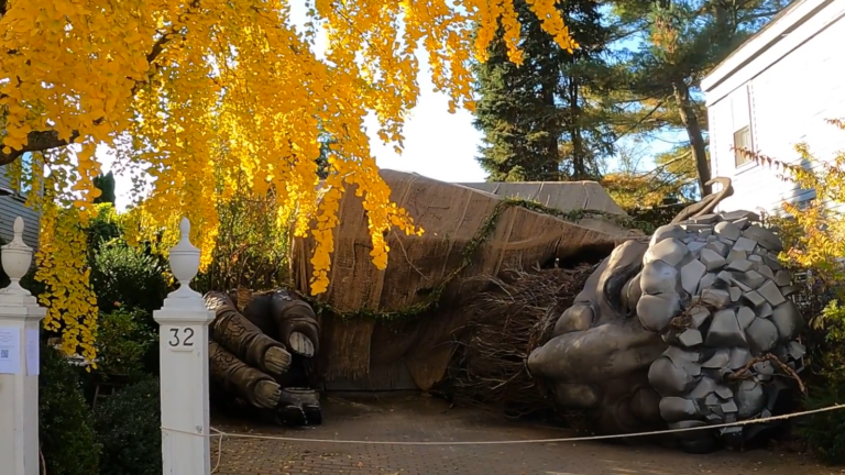 A giant troll statue on its side in a driveway with yellow fall leaves in the foreground.
