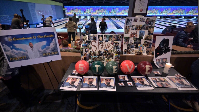 A vigil at a bowling alley in Malden includes photos of Steve Vozzella, a victim of the Lewiston mass shooting, and five bowling balls that spell out Steve's name using tape. Behind the vigil are several people and bowling lanes.