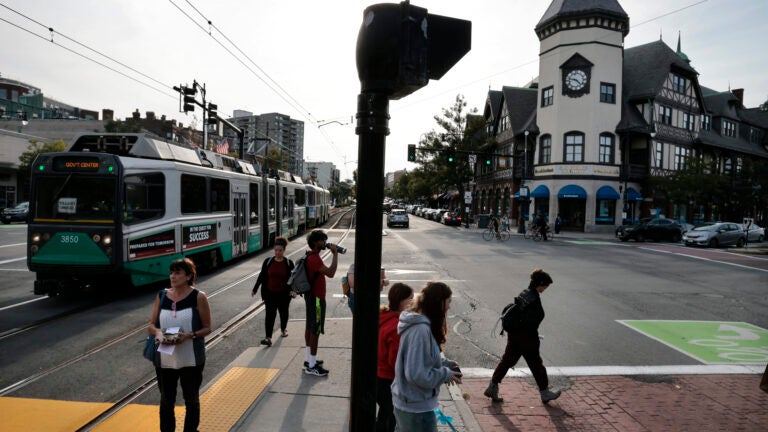 People walk in Coolidge Corner of Brookline as a MBTA Green Line car moves along the C Branch tracks.