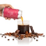 An image of a cold coffee can being poured into a glass, next to nuts and pieces of chocolate.