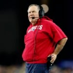 New England Patriots head coach Bill Belichick wearing a red New England Patriots hoodie with cutoff sleeves.