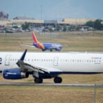 A Jet Blue jetliner taxis down a runway as a Southwest Airlines airliner takes off from Denver International Airport.