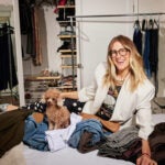 The stylist Chellie Carlson sits on a bed full of clothes in front of a closet and a handing rack. Her brown toy poodle is on the bed.