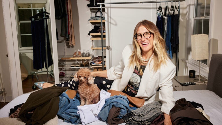 The stylist Chellie Carlson sits on a bed full of clothes in front of a closet and a handing rack. Her brown toy poodle is on the bed.