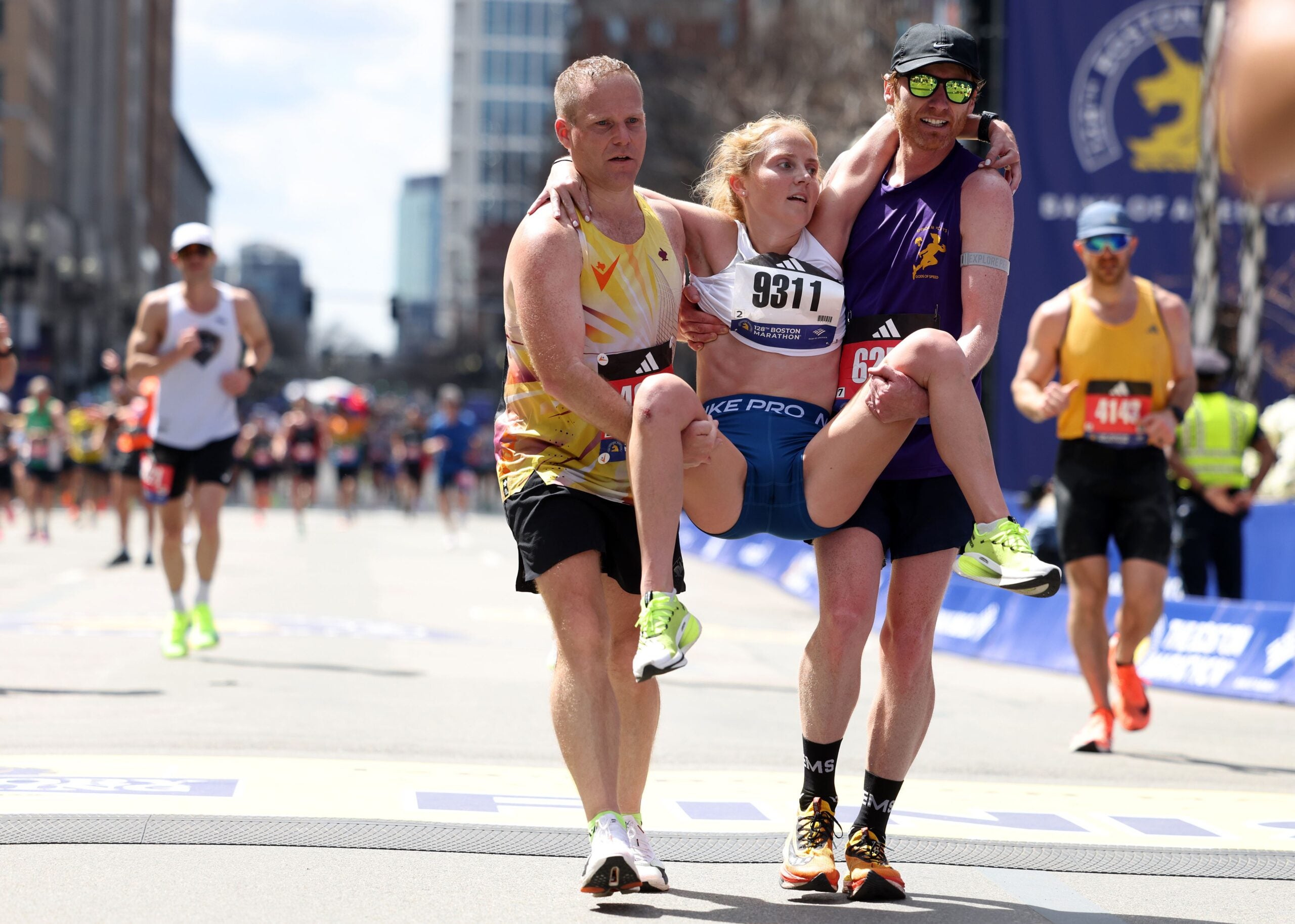 Merritt Bloom (C) viene aiutato a tagliare il traguardo da due compagni corridori durante la maratona di Boston di lunedì 15 aprile 2024. 