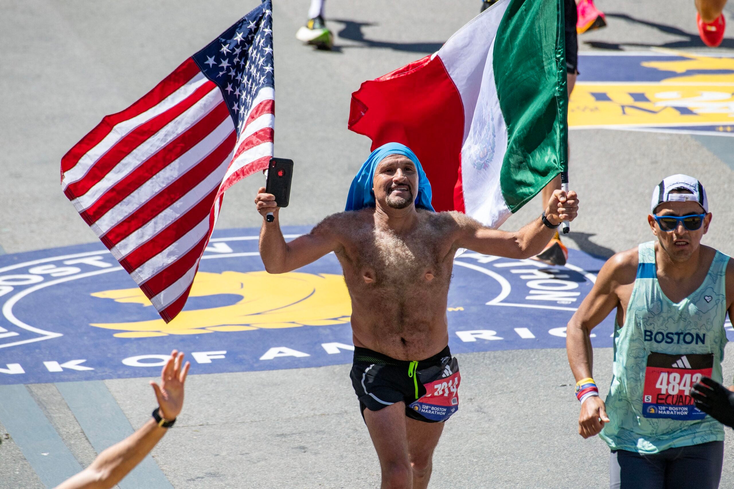 A runner carries an American flag and Mexican flag as he runs toward the finish line during the 128th Boston Marathon on April 15, 2024, in Boston, Massachusetts. The marathon includes around 30,000 athletes from 129 countries running the 26.2 miles from Hopkinton to Boston.