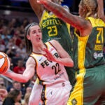 Indiana Fever guard Caitlin Clark passes the ball from under the basket while being defended by Seattle Storm guard Jordan Horston and center Mercedes Russell.