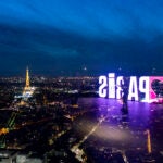 A visitor strolls past a "Paris" sign that is reflected in the glass wall of the Montparnasse tower observation deck as the Eiffel Tower stands in the background ahead of the 2024 Summer Olympics.