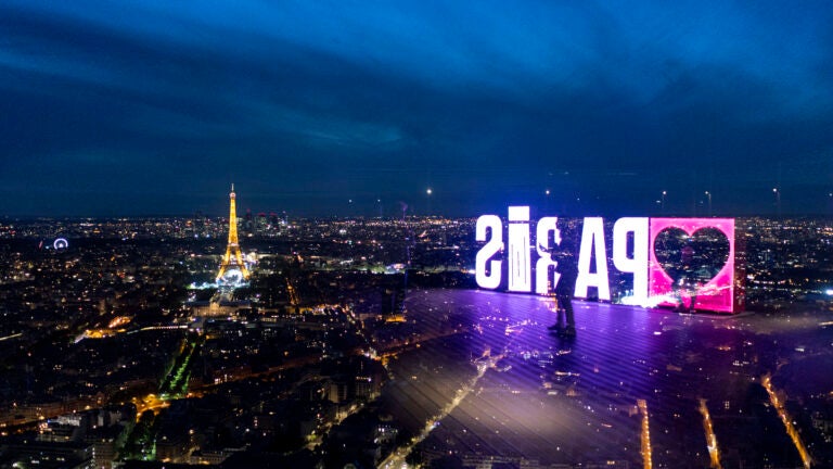 A visitor strolls past a "Paris" sign that is reflected in the glass wall of the Montparnasse tower observation deck as the Eiffel Tower stands in the background ahead of the 2024 Summer Olympics.