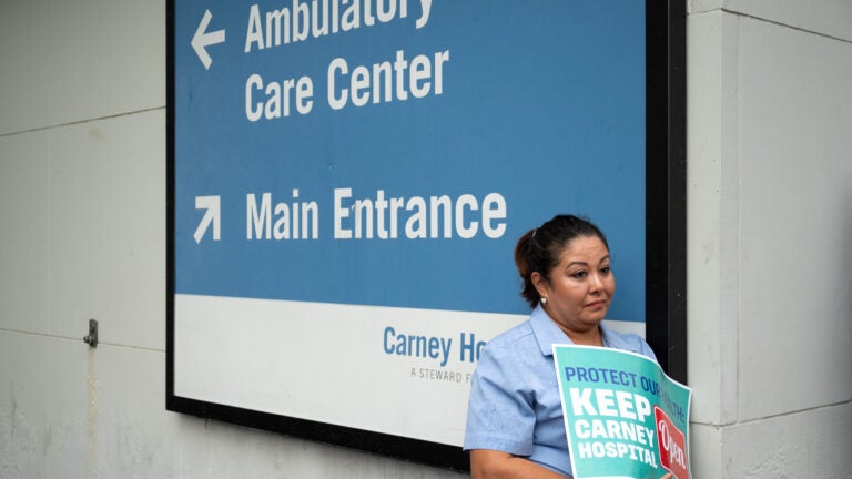 A person holds a sign reading “protect our health: Keep Carney Hospital Open” while standing near a sign at Carney Hospital in Dorchester.