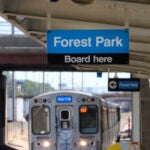 A security camera and speaker hang from the ceiling of the Chicago Transit Authority Harlem Ave. station as a Blue Line train enters the station heading West to the Forest Park, Ill., station as two pedestrians walk toward the station.