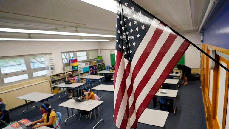 Una bandera estadounidense cuelga en un salón de clases donde los estudiantes trabajan en computadoras portátiles.