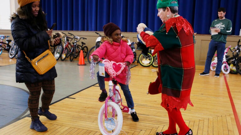 Charli Jenkins mounts her new bicycle as Lynne Smith, 74, picks up a stuffed animal that fell on the ground during a toy drive in Dorchester on Christmas Eve. Action for Boston Community Development is struggling to get enough toy donations for its toy drive to meet the need of families this year.
