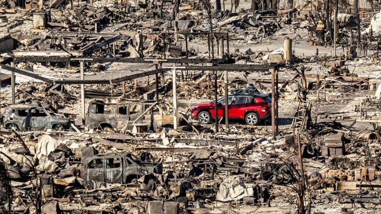 A car drives past homes and vehicles destroyed by the Palisades Fire.