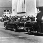 Secret servicemen standing on running boards follow the presidential limousine carrying President John F. Kennedy.